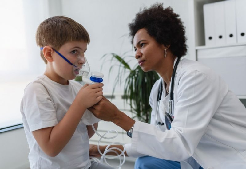 6022403270c5c Woman African American doctor general practitioner helping child to put nebulizer inhaler face mask. Asthma treatment for children.